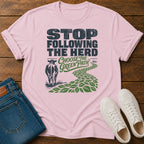 A light pink graphic t-shirt laid flat on a wooden surface, styled with folded blue jeans and white sneakers. The t-shirt features a graphic with the text 'STOP FOLLOWING THE HERD' and 'CHOOSE THE GREEN PATH' alongside an illustration of a cow and a green leafy path.