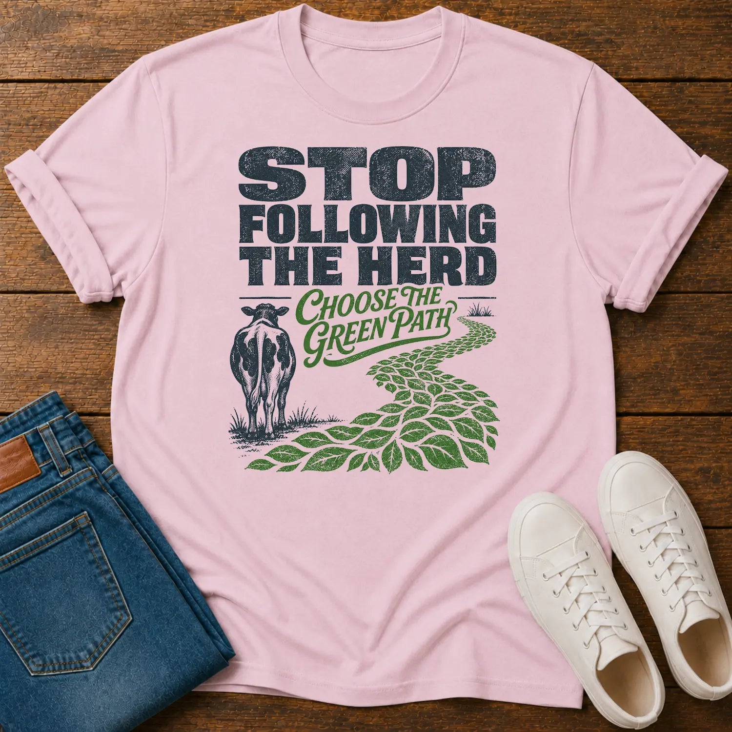 A light pink graphic t-shirt laid flat on a wooden surface, styled with folded blue jeans and white sneakers. The t-shirt features a graphic with the text 'STOP FOLLOWING THE HERD' and 'CHOOSE THE GREEN PATH' alongside an illustration of a cow and a green leafy path.