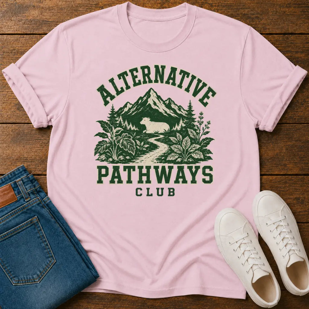 A light pink short-sleeve graphic T-shirt laid flat on a wooden surface, styled with folded blue jeans and white sneakers. The shirt has a dark green printed design reading 'ALTERNATIVE PATHWAYS CLUB' with mountains, trees, and a path.