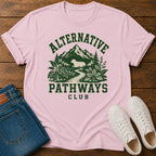 A light pink short-sleeve graphic T-shirt laid flat on a wooden surface, styled with folded blue jeans and white sneakers. The shirt has a dark green printed design reading 'ALTERNATIVE PATHWAYS CLUB' with mountains, trees, and a path.