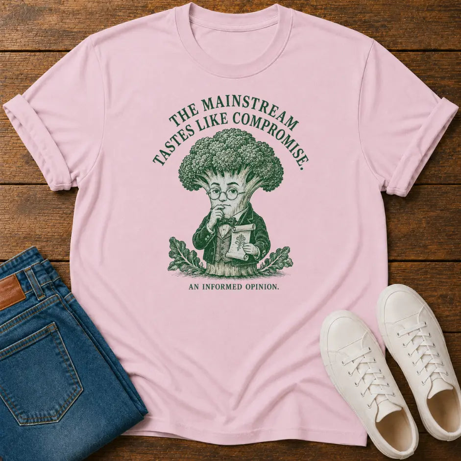 A light pink graphic t-shirt laid flat on a wooden surface, styled with folded blue jeans and white sneakers. The t-shirt features a dark green graphic of a broccoli-headed character with the text 'THE MAINSTREAM TASTES LIKE COMPROMISE.
