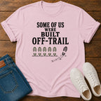 A light pink graphic t-shirt laid flat on a dark wood background, styled with folded blue jeans and white sneakers. The t-shirt features the black printed text 'SOME OF US WERE BUILT OFF-TRAIL' above a line of green camouflage bootprints and a black footprint trail graphic.