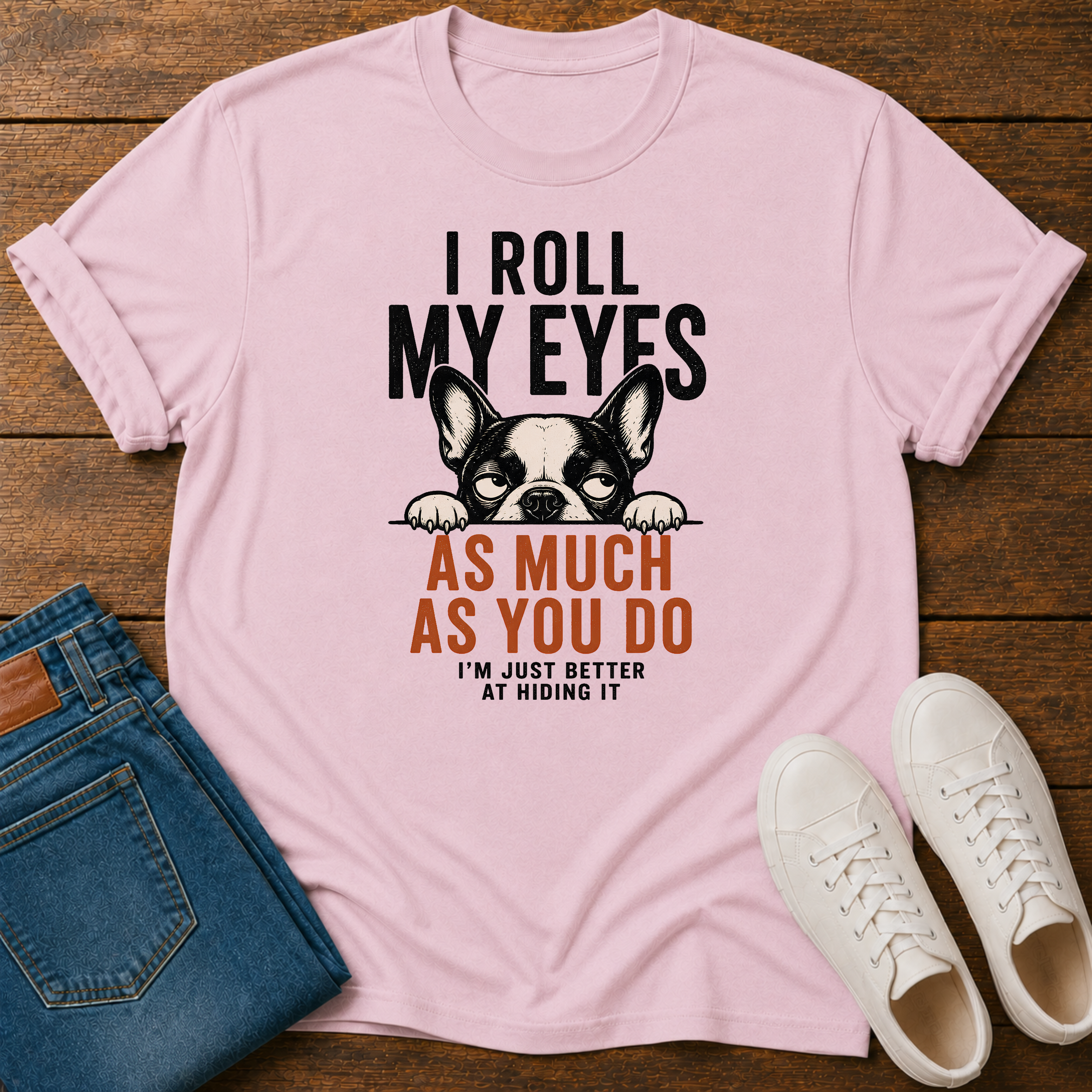 A light pink graphic t-shirt laid flat on a dark wood background, styled with folded blue jeans and white sneakers. The t-shirt features a black and white French Bulldog graphic with the text 'I ROLL MY EYES AS MUCH AS YOU DO I'M JUST BETTER AT HIDING IT'.