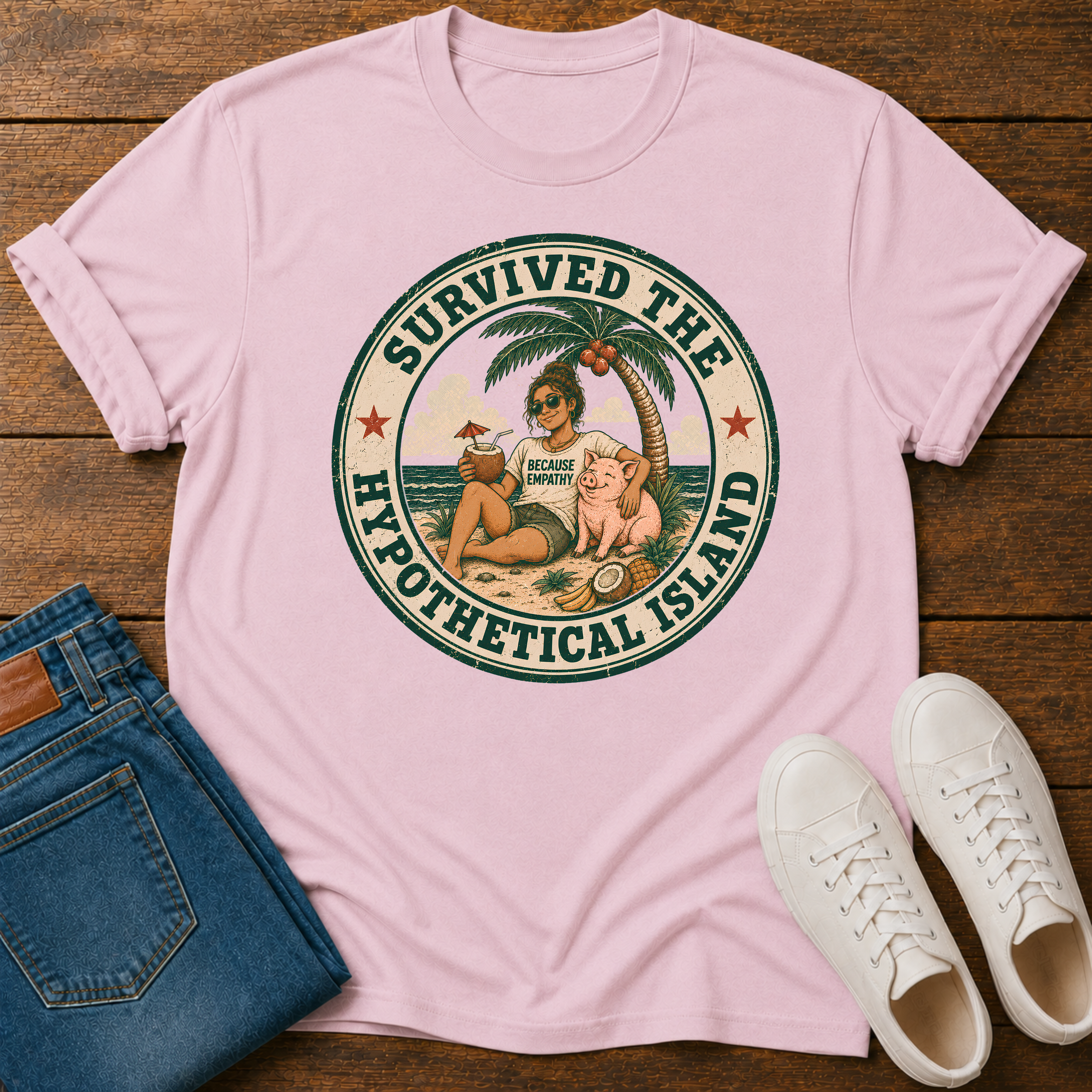 A light pink short-sleeve graphic T-shirt laid flat on a wooden surface, styled with folded blue jeans and white sneakers. The shirt features a circular retro beach illustration with the text 'SURVIVED THE HYPOTHETICAL ISLAND' and a small 'Tropical Island' logo in the center.