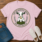 A light pink crewneck t-shirt laid flat on a dark wood background, styled with a pair of blue jeans and white sneakers. The t-shirt features a circular graphic of a black-and-white cow holding a gold bell, with the text 'FOR THE RIGHTFUL OWNER RESERVED NOT YOUR BOTTE' in a vintage license-style design.