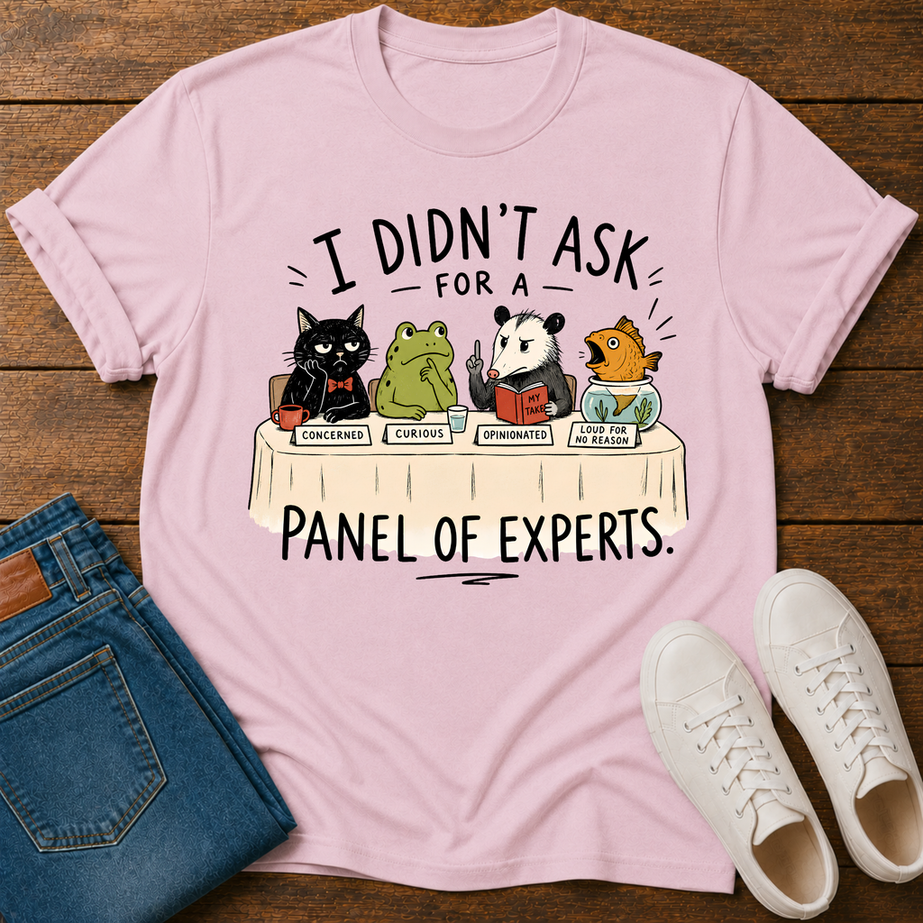 A light pink graphic t-shirt laid flat on a dark wood background, styled with blue jeans and white sneakers. The t-shirt features a cartoon graphic of four animals (a black cat, a green frog, a panda, and a fish) sitting at a table labeled 'PANEL OF EXPERTS' with humorous labels for their responses.