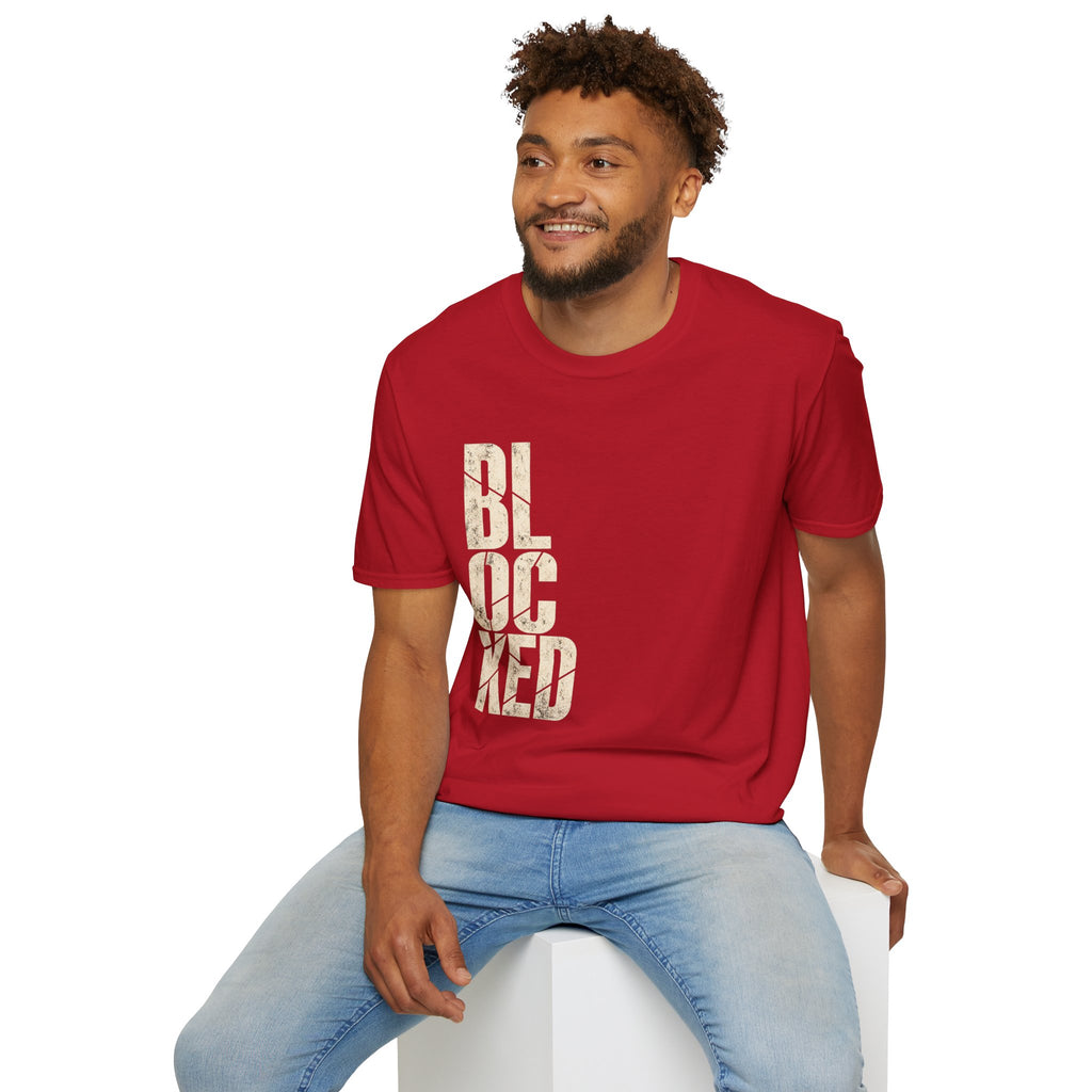 A man wearing a red crewneck t-shirt with the word 'BLOCKED' printed vertically in distressed beige block letters, paired with light blue jeans, sitting on a white block against a plain white background.
