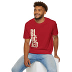 A man wearing a red crewneck t-shirt with the word 'BLOCKED' printed vertically in distressed beige block letters, paired with light blue jeans, sitting on a white block against a plain white background.