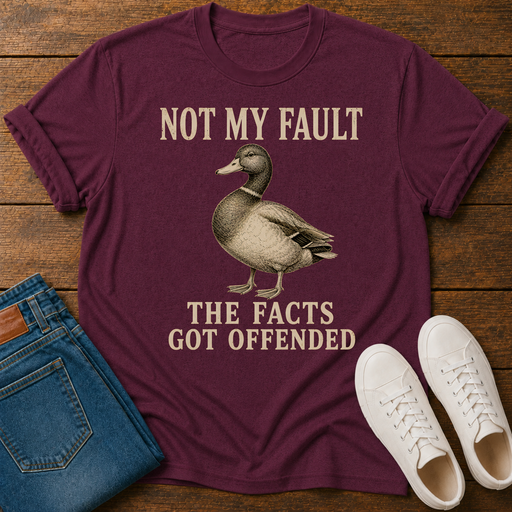 A maroon short-sleeve graphic T-shirt laid flat on a wooden surface, styled with blue jeans and white sneakers. The shirt has a vintage-style duck illustration and the text 'NOT MY FAULT THE FACTS GOT OFFENDED' printed in a light tan color.