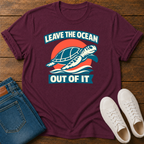 A maroon short-sleeve graphic T-shirt laid flat on a wooden surface, styled with folded blue jeans and white sneakers. The shirt has a large turtle-and-sunset print with the text “LEAVE THE OCEAN OUT OF IT.