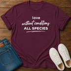 A maroon short-sleeve graphic T-shirt laid flat on a wooden surface, styled with folded blue jeans and a pair of white sneakers. The shirt has white text reading 'love without conditions ALL SPECIES'.
