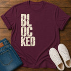 A maroon short-sleeve t-shirt with the word 'BLOCKED' printed vertically in distressed off-white block letters, styled in a flat lay on a dark wood background alongside folded blue jeans and a pair of white sneakers.