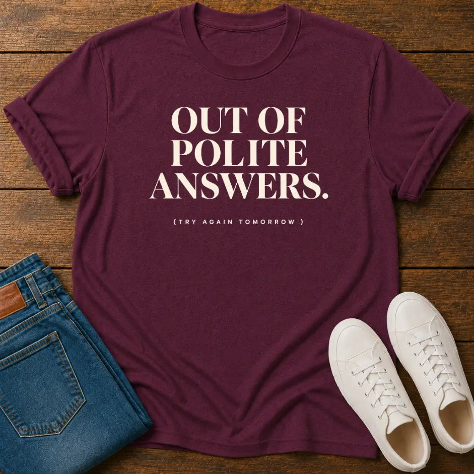 A maroon short-sleeve graphic T-shirt laid flat on a wooden surface, styled with folded blue jeans and a pair of white sneakers. The shirt has large white text reading “OUT OF POLITE ANSWERS.