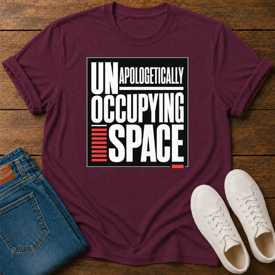 A maroon short-sleeve graphic T-shirt laid flat on a wooden surface, styled with folded blue jeans and white sneakers. The shirt has a large front print reading “UNAPOLOGETHICALLY OCCUPING SPACE” in white text with a red striped accent.