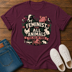 A maroon short-sleeve graphic T-shirt laid flat on a wooden surface, styled with folded blue jeans and white sneakers. The shirt features a large front print with illustrated farm animals and the text 'FEMINIST FOR ALL ANIMALS YES, ALL OF THEM.