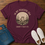 A maroon short-sleeve graphic T-shirt laid flat on a wooden surface, styled with folded blue jeans and white sneakers. The shirt has a circular vintage-style frog illustration in beige and text reading “IN MY PEACEFUL ERA (DON’T TEST IT)”.