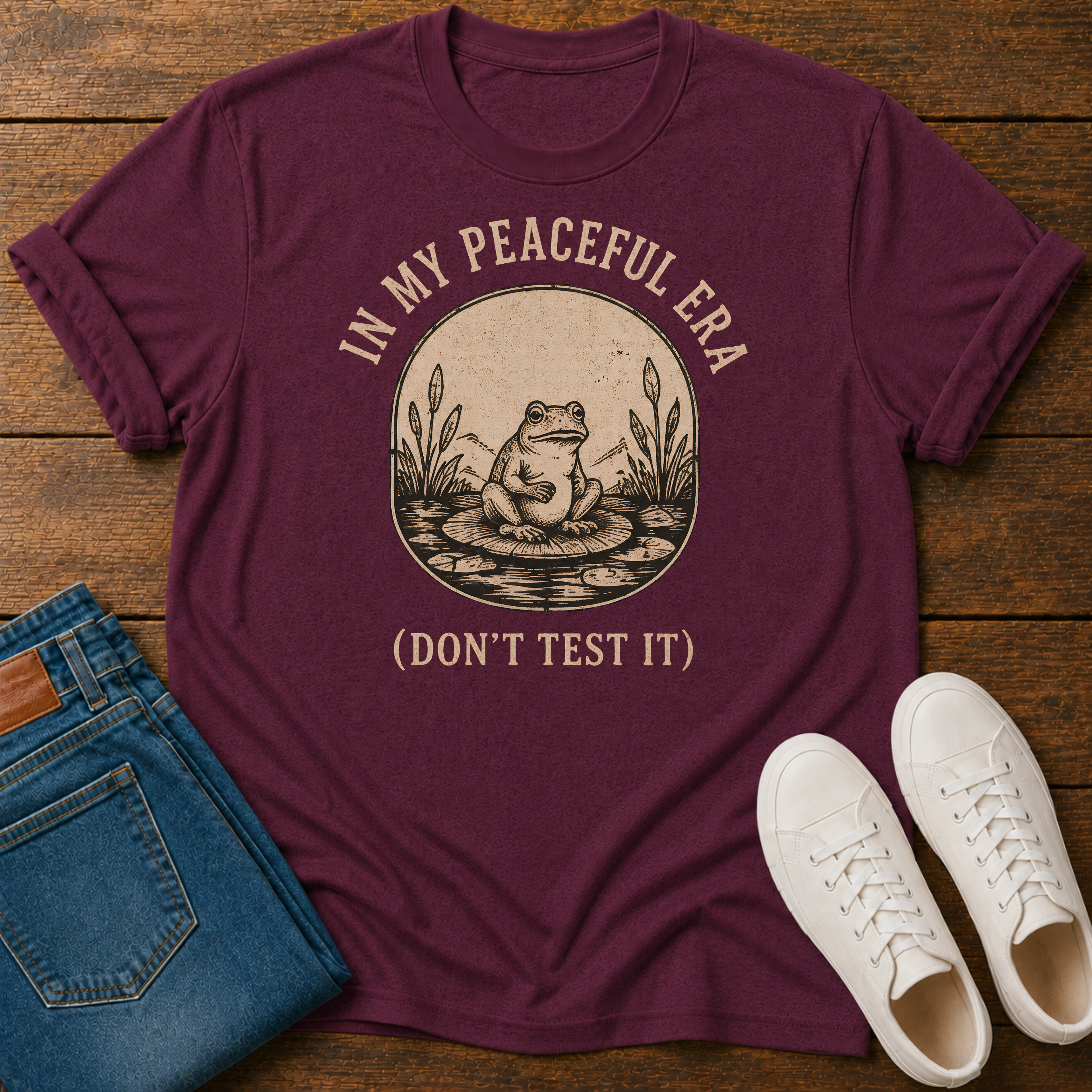 A maroon short-sleeve graphic T-shirt laid flat on a wooden surface, styled with folded blue jeans and white sneakers. The shirt has a circular vintage-style frog illustration in beige and text reading “IN MY PEACEFUL ERA (DON’T TEST IT)”.