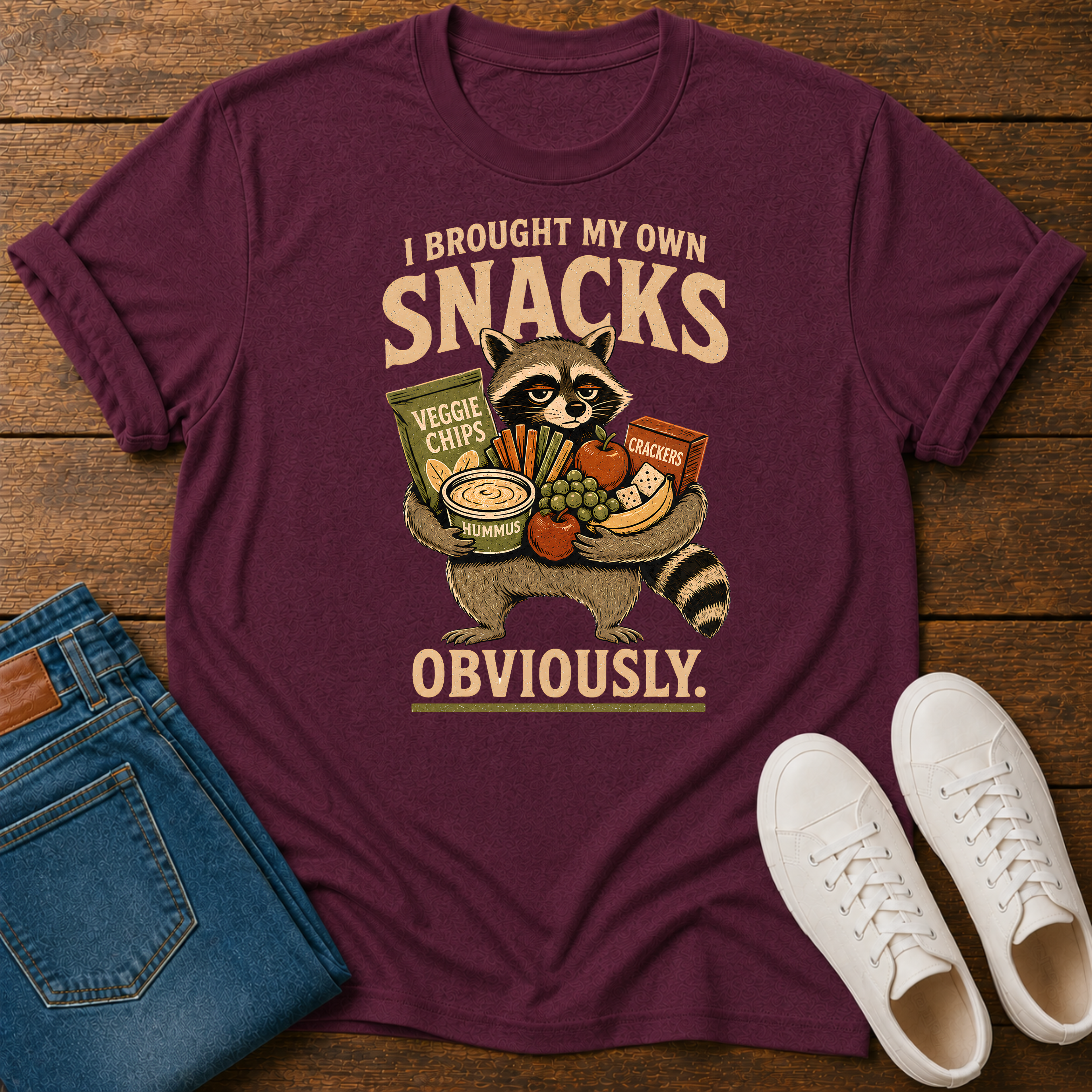 A maroon short-sleeve graphic T-shirt laid flat on a wooden surface, styled with folded blue jeans and white sneakers. The shirt features a large front print of a raccoon holding snacks with the text 'I BROUGHT MY OWN SNACKS OBVIOUSLY.