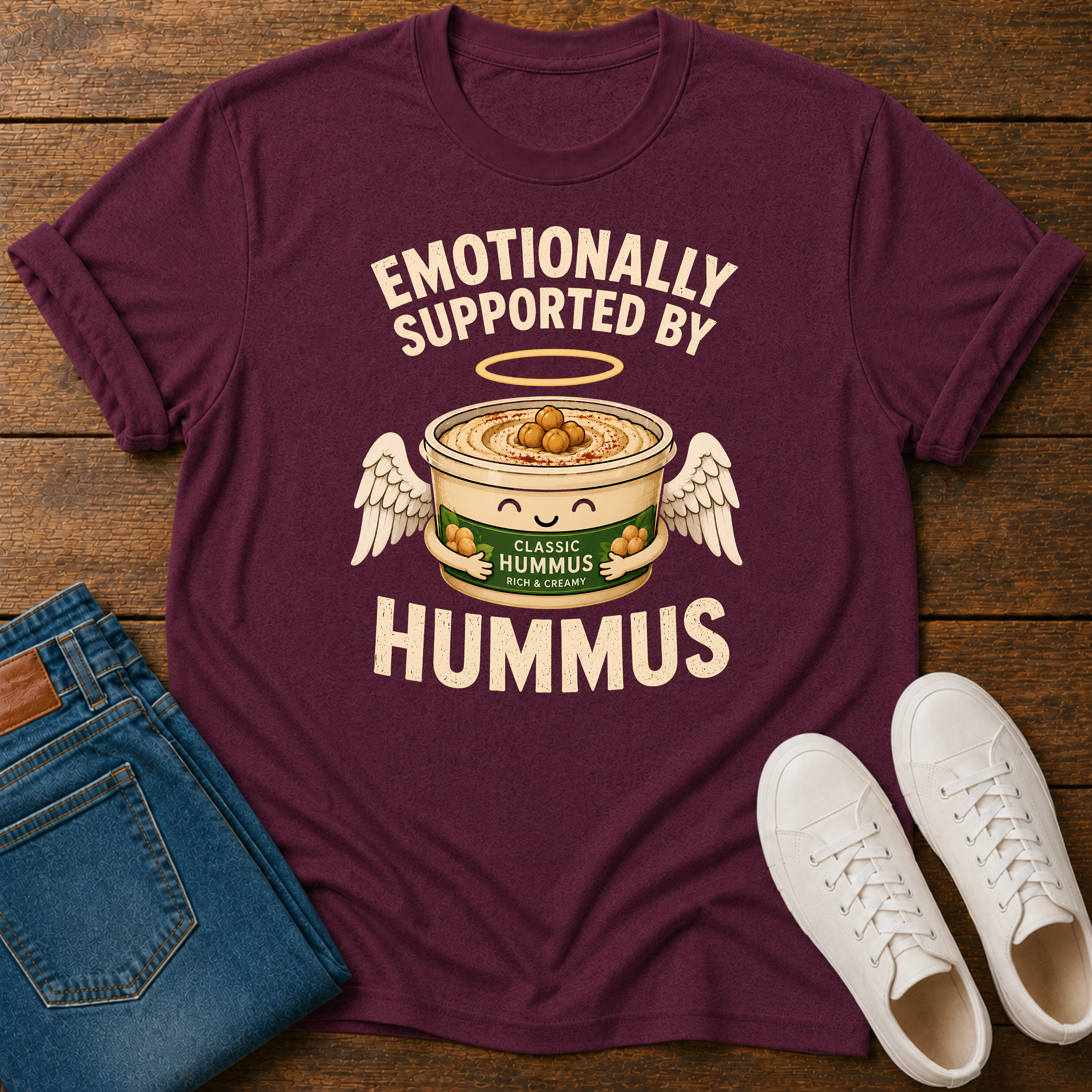 A maroon short-sleeve graphic T-shirt laid flat on a wooden surface, styled with folded blue jeans and white sneakers. The shirt has a large cream graphic and text reading “EMOTIONALLY SUPPORTED BY HUMMUS,” featuring a cartoon hummus bowl with wings and olives.