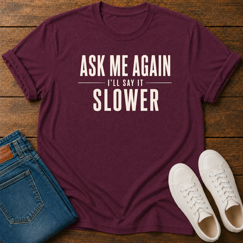 A maroon short-sleeve graphic T-shirt laid flat on a wooden surface, styled with folded blue jeans and white sneakers. The shirt has large white text reading “ASK ME AGAIN I'LL SAY IT SLOWER.