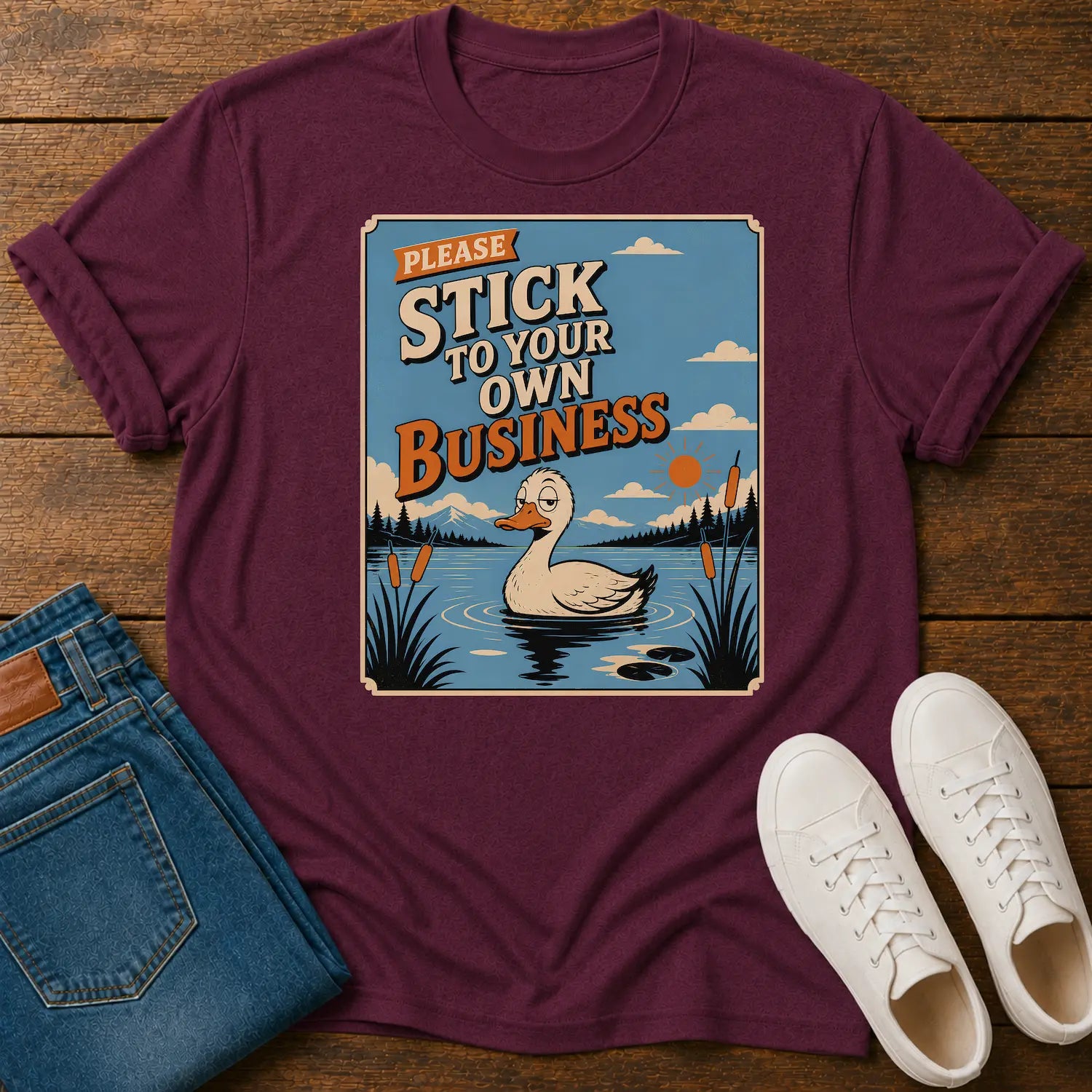 A maroon short-sleeve graphic T-shirt laid flat on a wooden surface, styled with folded blue jeans and white sneakers. The shirt features a square vintage-style print of a duck in a pond with the text 'PLEASE STICK TO YOUR OWN BUSINESS.