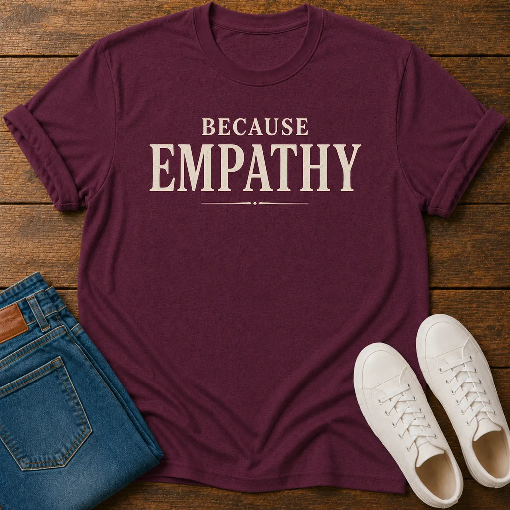A maroon short-sleeve graphic T-shirt laid flat on a wooden surface, printed with the words 'BECAUSE EMPATHY' in cream. Folded blue jeans and a pair of white sneakers are placed beside it as styling props.