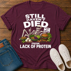 Maroon t-shirt with skeleton graphic and protein text on a wooden surface with jeans and sneakers.