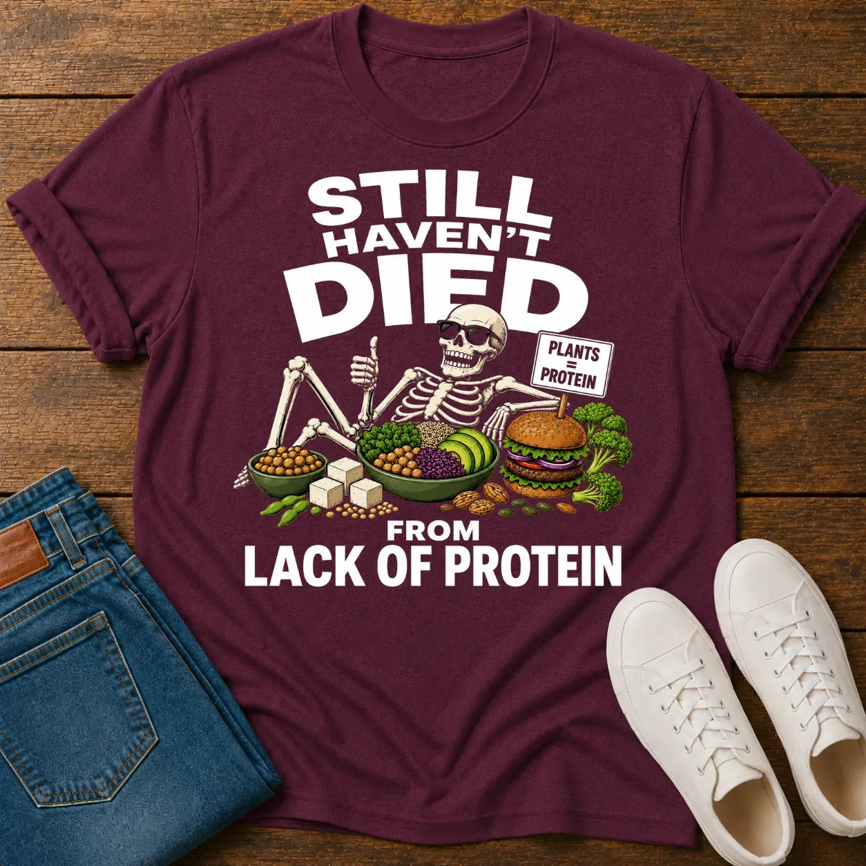 Maroon t-shirt with skeleton graphic and protein text on a wooden surface with jeans and sneakers.