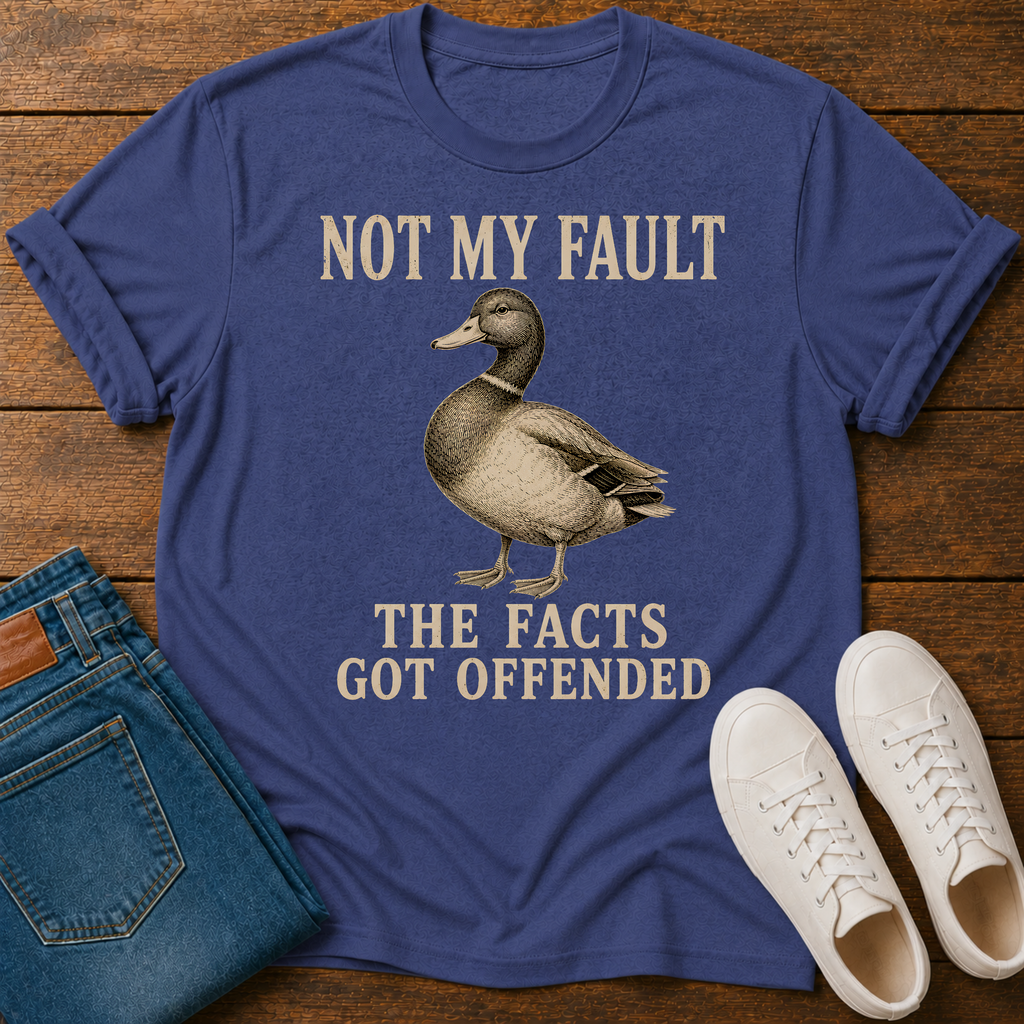 A blue graphic t-shirt laid flat on a wooden surface, styled with folded blue jeans, white sneakers, and a small white cup. The t-shirt features a vintage-style duck illustration and the text 'NOT MY FAULT THE FACTS GOT OFFENDED' in beige.