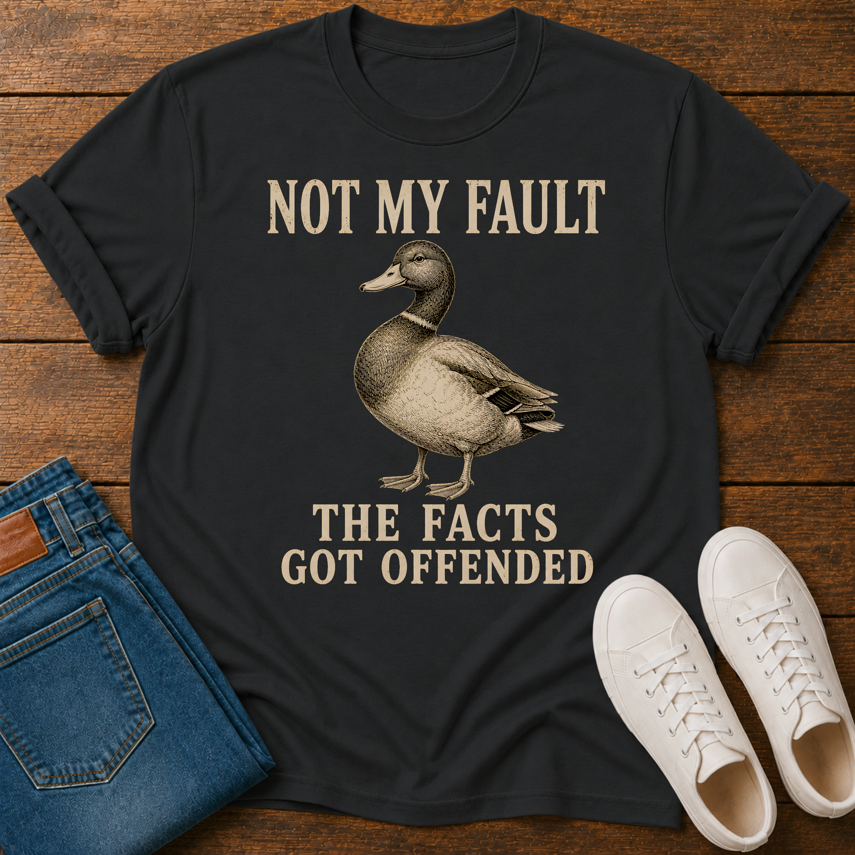 A black graphic t-shirt featuring a duck illustration and the text 'NOT MY FAULT THE FACTS GOT OFFENDED' in beige, styled in a flat lay with blue jeans and white sneakers on a wooden background.