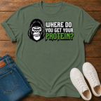 A military green short-sleeve graphic T-shirt laid flat on a wooden surface, styled with folded blue jeans and white sneakers. The shirt features a large gorilla skull design with leaf details, crossed plant stems, and the text ''where do you get your protein from?"
