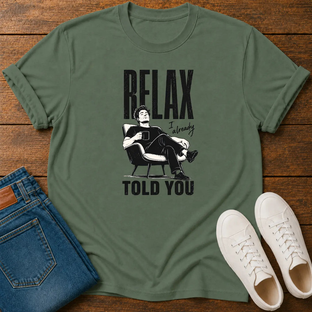 An olive green graphic t-shirt featuring a black and white illustration of Elon Musk sitting in a chair with the text 'RELAX I'm ready TOLD YOU', styled in a flat lay on a wooden background with blue jeans and white sneakers.