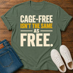 An military green graphic t-shirt laid flat on a wooden surface, styled with folded blue jeans and white sneakers. The t-shirt features the text 'CAGE-FREE ISN'T THE SAME AS FREE.