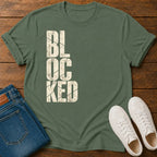 An military green crewneck t-shirt with a distressed cream-colored 'BLOCKED' graphic printed vertically on the front. The t-shirt is styled in a flat-lay arrangement on a dark wood background, accompanied by folded blue jeans and a pair of white sneakers.