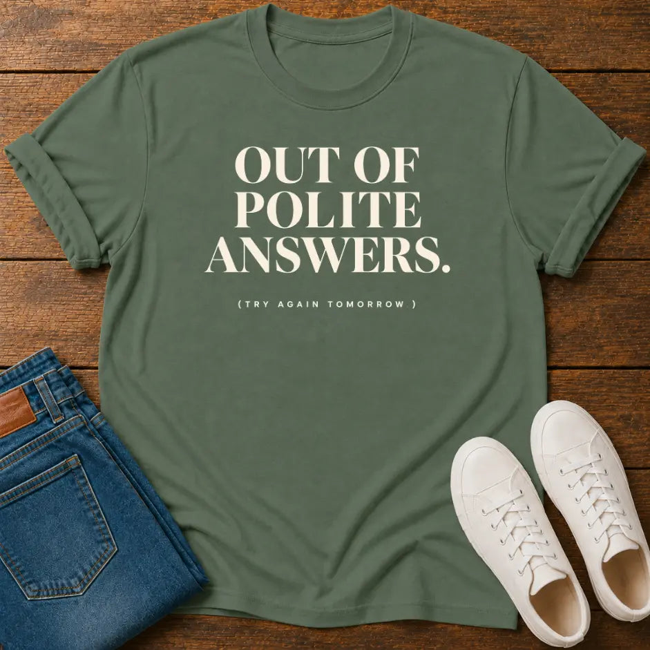 An olive green graphic t-shirt with the text 'OUT OF POLITE ANSWERS. (TRY AGAIN TOMORROW)' printed in white, styled in a flat-lay on a wooden background with blue jeans and white sneakers.