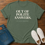 An olive green graphic t-shirt with the text 'OUT OF POLITE ANSWERS. (TRY AGAIN TOMORROW)' printed in white, styled in a flat-lay on a wooden background with blue jeans and white sneakers.
