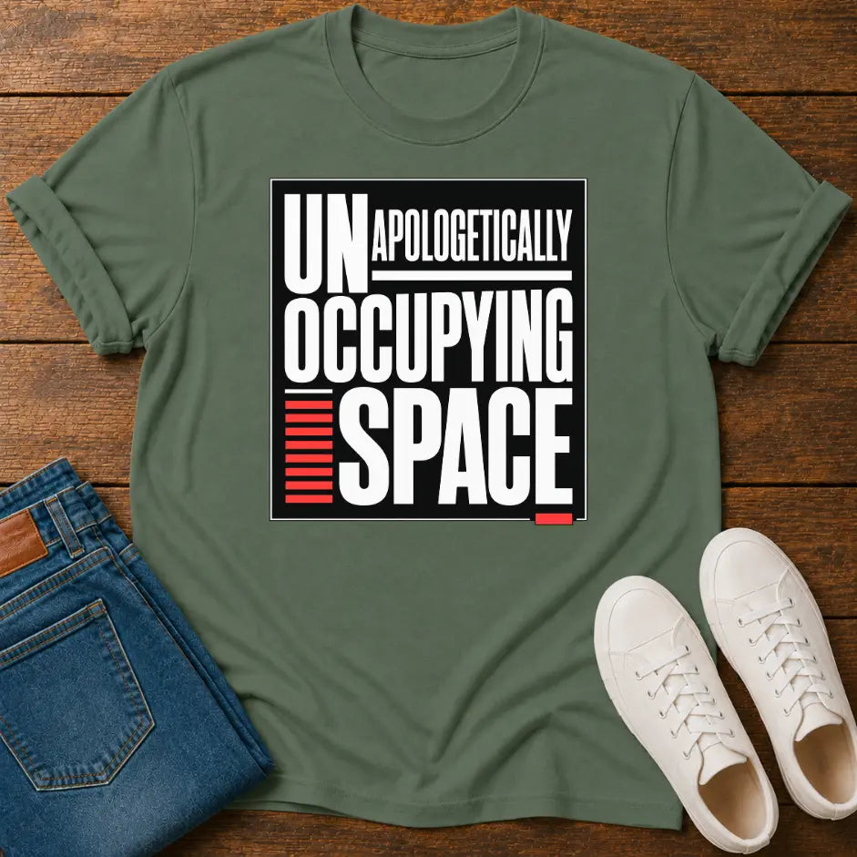 An olive green graphic t-shirt laid flat on a wooden surface, styled with folded blue jeans and a pair of white sneakers. The t-shirt features a bold black and white graphic with red accents that reads 'UNAPOLOGETICALLY OCCUPYING SPACE'.