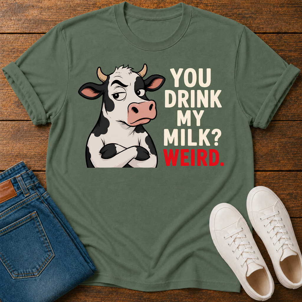 An olive green graphic t-shirt laid flat on a wooden surface, styled with folded blue jeans and white sneakers. The t-shirt features a cartoon cow with crossed arms next to the text 'YOU DRINK MY MILK? WEIRD.