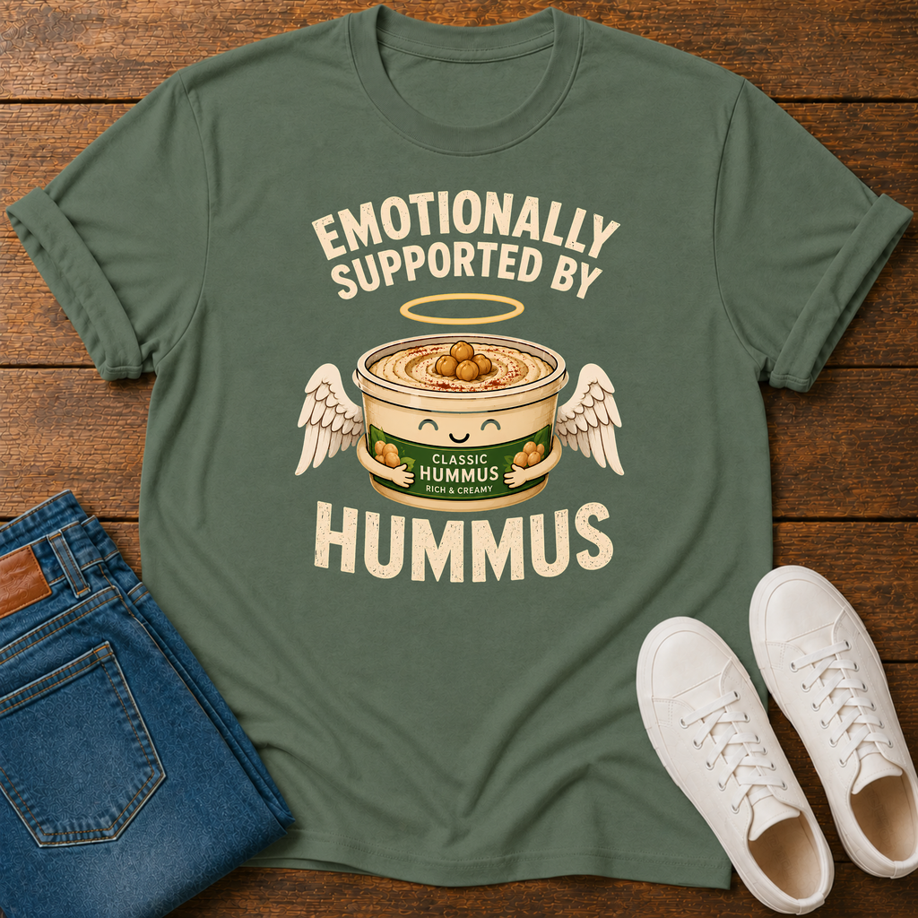 An military green graphic t-shirt laid flat on a wooden surface, styled with folded blue jeans and white sneakers. The t-shirt features a graphic of a can of hummus with angel wings and a halo, with the text 'EMOTIONALLY SUPPORTED BY HUMMUS'.