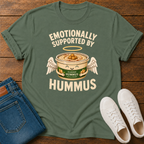 An military green graphic t-shirt laid flat on a wooden surface, styled with folded blue jeans and white sneakers. The t-shirt features a graphic of a can of hummus with angel wings and a halo, with the text 'EMOTIONALLY SUPPORTED BY HUMMUS'.