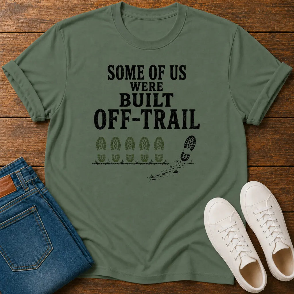 An olive green graphic t-shirt laid flat on a wooden surface, styled with folded blue jeans and a pair of white sneakers. The t-shirt features the black printed text 'SOME OF US WERE BUILT OFF-TRAIL' above a trail footprint graphic.