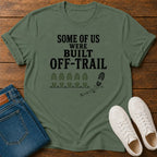 An olive green graphic t-shirt laid flat on a wooden surface, styled with folded blue jeans and a pair of white sneakers. The t-shirt features the black printed text 'SOME OF US WERE BUILT OFF-TRAIL' above a trail footprint graphic.