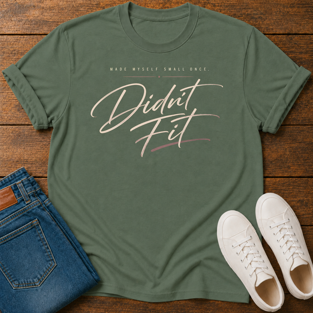 A military green graphic t-shirt laid flat on a wooden surface, styled with folded blue jeans and white sneakers. The t-shirt features the text 'MADE MYSELF SMALL ONCE. DIDN'T FIT'
