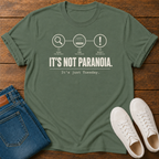 A military green short-sleeve graphic T-shirt laid flat on a wooden surface, styled with folded blue jeans and white sneakers.  text 'it's not paranoia it's a tuesday'