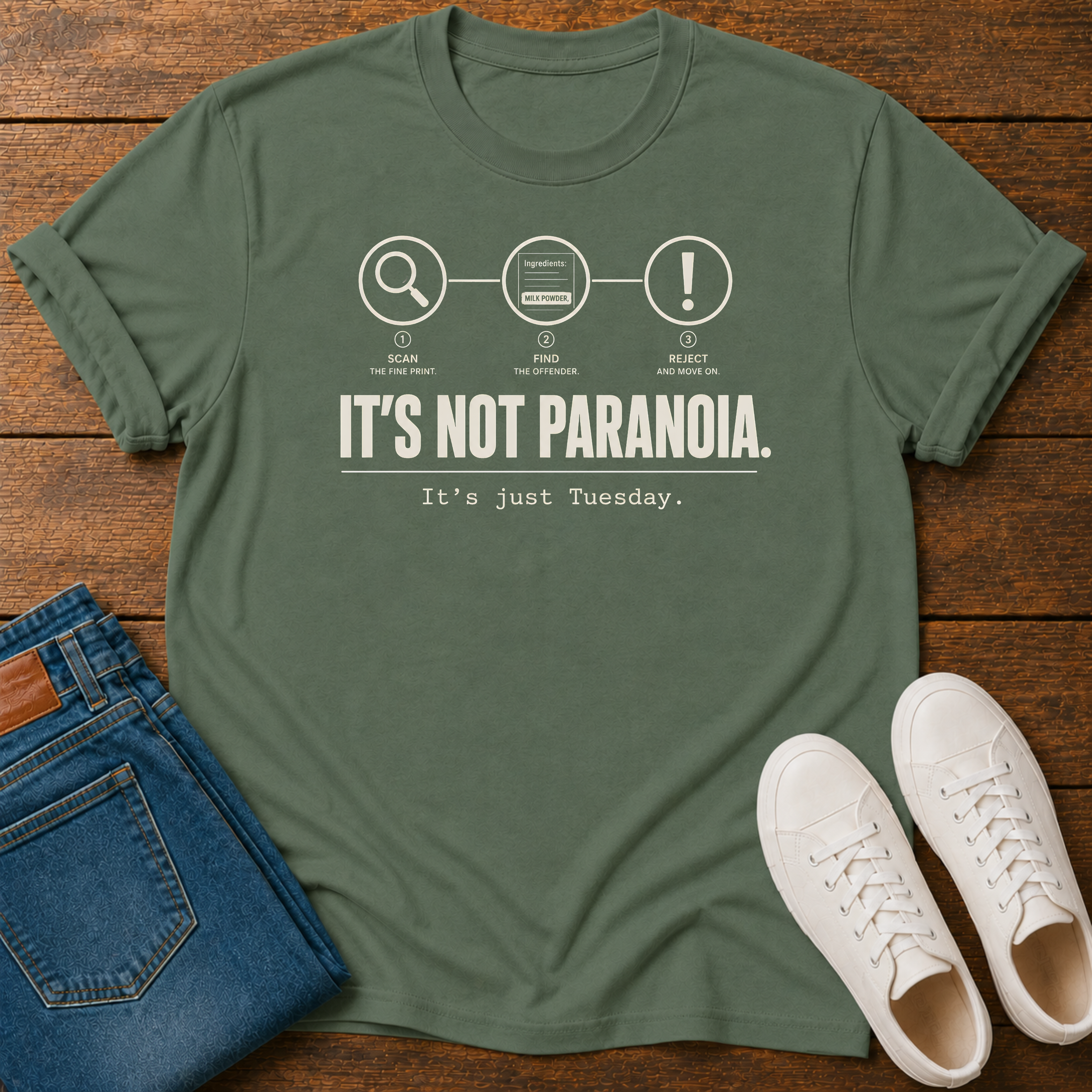 A military green short-sleeve graphic T-shirt laid flat on a wooden surface, styled with folded blue jeans and white sneakers.  text 'it's not paranoia it's a tuesday'