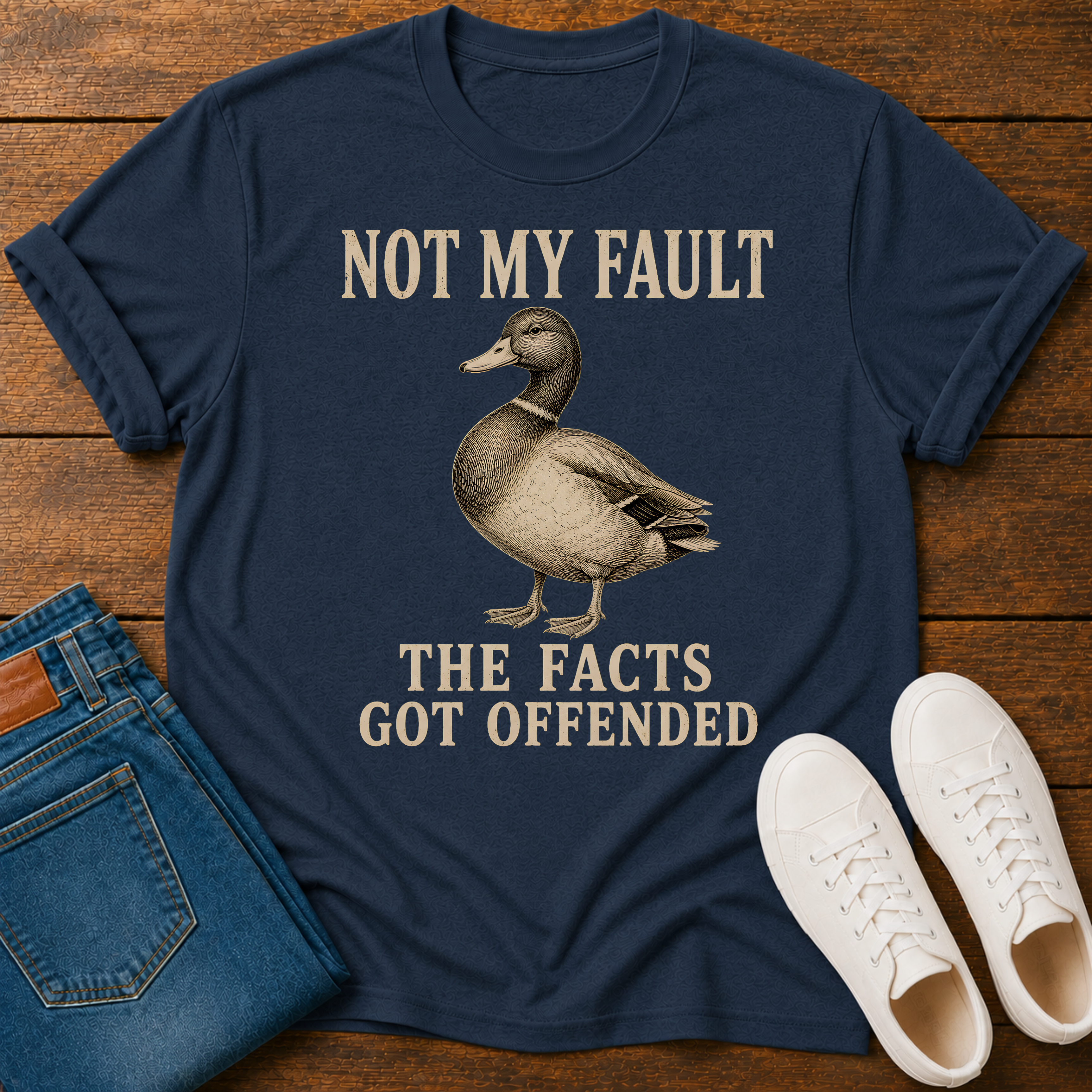 A navy blue graphic t-shirt laid flat on a wooden surface, styled with folded blue jeans, white sneakers, and a duck illustration with the text 'NOT MY FAULT THE FACTS GOT OFFENDED'.