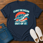 A navy blue graphic t-shirt laid flat on a wooden surface, styled with folded blue jeans and white sneakers. The t-shirt features a retro-style graphic of a sea turtle swimming in front of a red-orange sun, with the text 'LEAVE THE OCEAN OUT OF IT'.