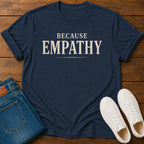 A navy blue graphic t-shirt with the words 'BECAUSE EMPATHY' printed in cream, styled in a flat lay on a wooden background with folded blue jeans and white sneakers.
