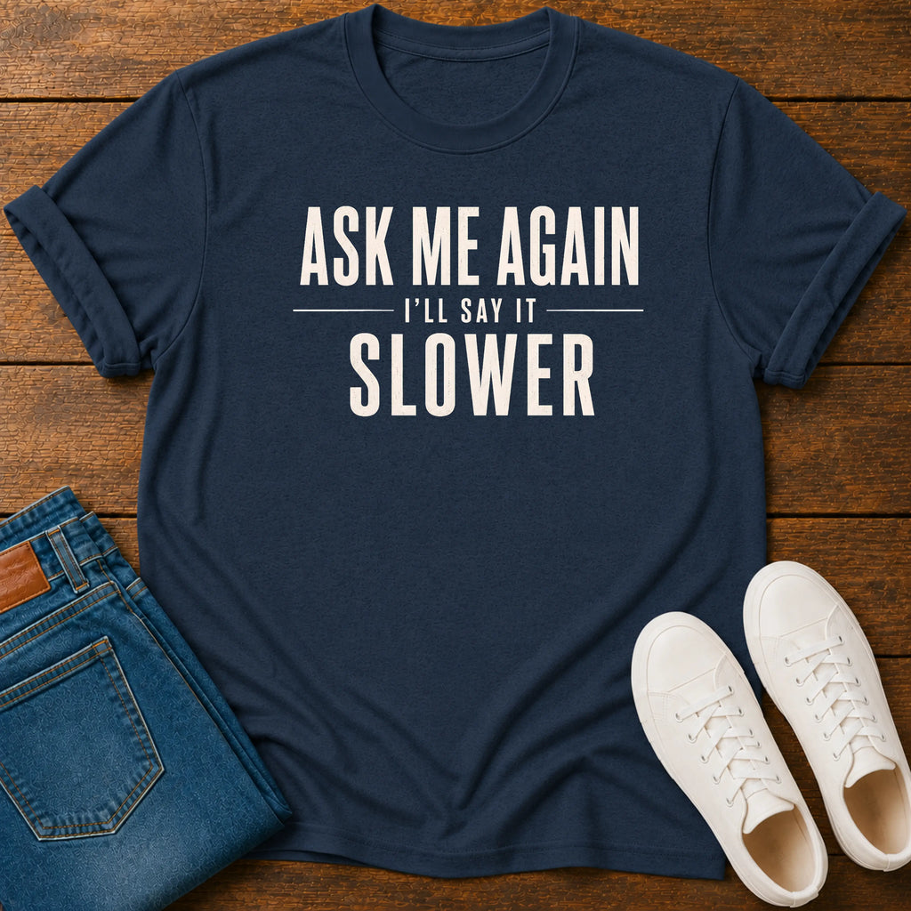 A navy blue graphic t-shirt with the text 'ASK ME AGAIN I'LL SAY IT SLOWER' in white, styled in a flat lay on a wooden background with folded blue jeans and white sneakers.