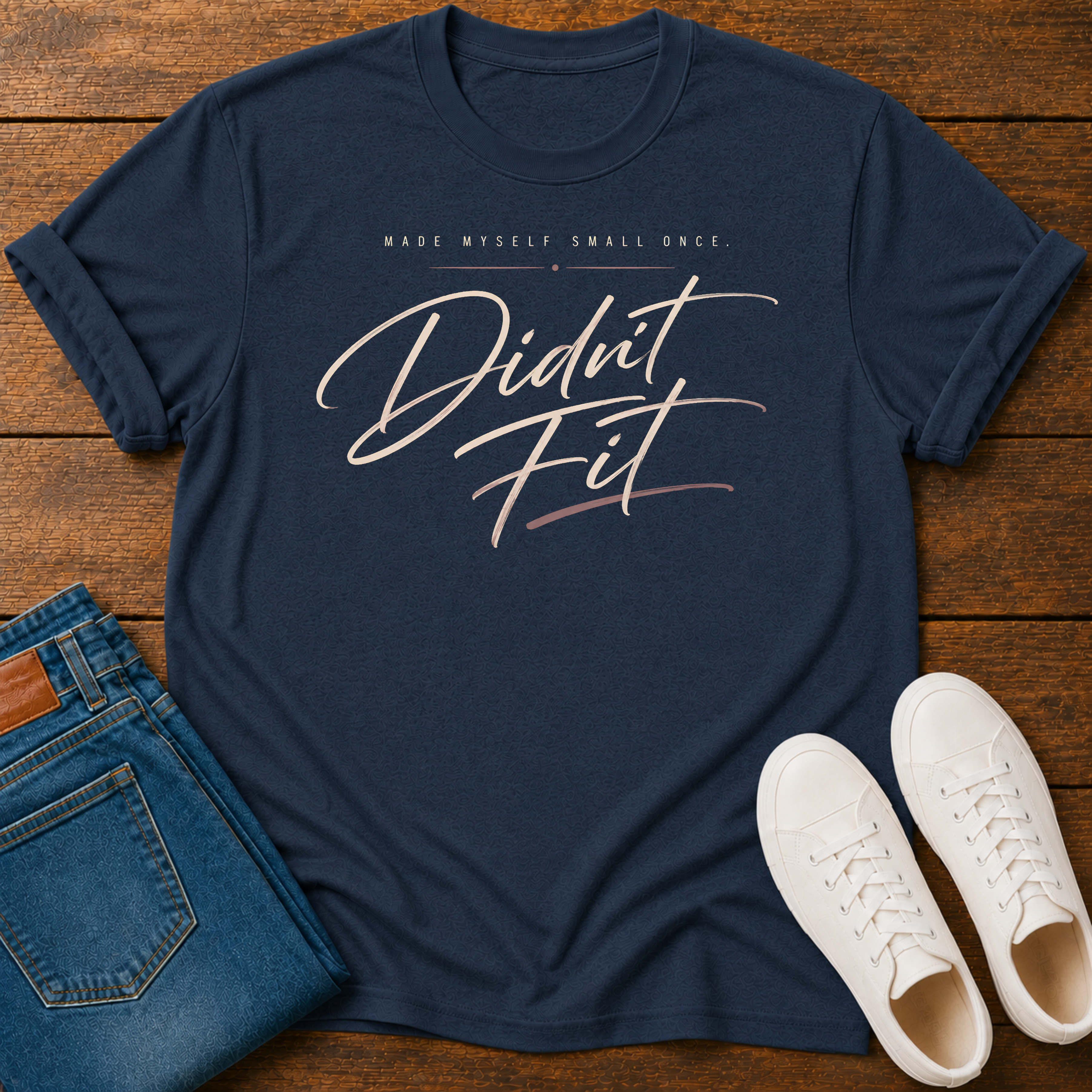 A navy blue graphic t-shirt laid flat on a wooden surface, styled with folded blue jeans and white sneakers. The t-shirt features the text 'MADE MYSELF SMALL ONCE. DIDN'T FIT'